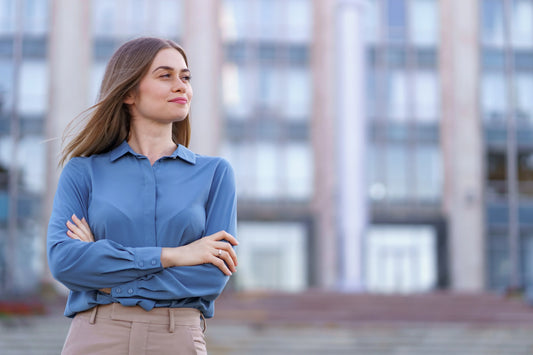 Confident young businesswoman in blue blouse with arms crossed standing outdoors by modern office building
