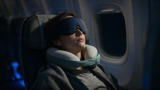 Passenger asleep in an airplane window seat wearing a navy contoured DreamShade sleep mask and a DreamElite travel pillow neck massager