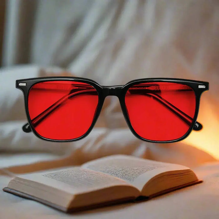Red lensed blue-light blocking glasses with black frame resting above an open book on cozy bedding